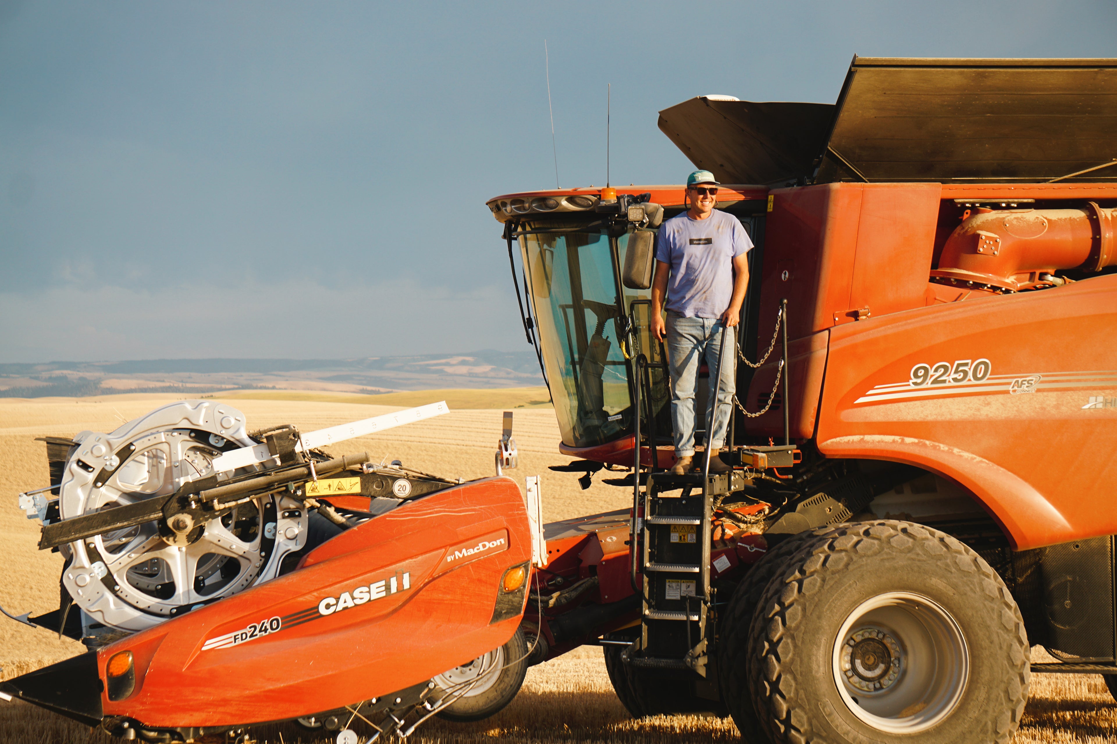 Man standing on a large Case combine in a field of wheat with mountains and sky in the background. 